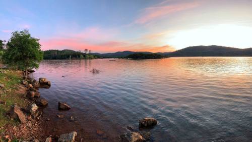 A serene lake at sunset, with calm waters reflecting colorful skies and surrounded by trees and hills.