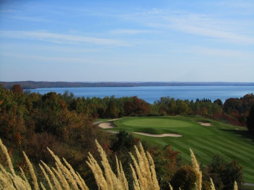 A scenic view of a golf course with rolling greens, surrounded by colorful autumn foliage and a calm lake in the background.