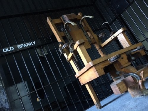 A wooden electric chair, labeled "OLD SPARKY," inside a barred cell, casting shadows on the floor.