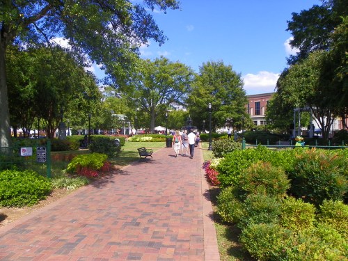 A sunny park pathway lined with greenery and flowers, with people walking and buildings in the background.