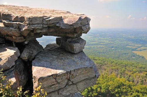 A rocky outcrop with a natural arch overlooks a lush green valley and distant hills under a clear blue sky.