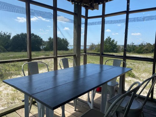 A wooden table surrounded by chairs in a screened-in area, with a view of greenery and blue sky outside.