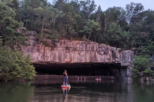 A person stands on a paddleboard in calm water near a rocky cave surrounded by lush greenery.