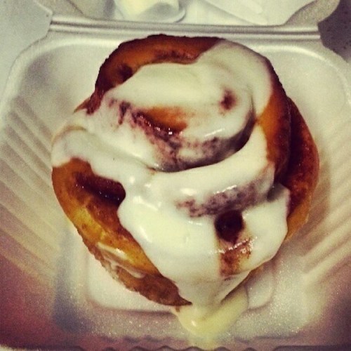 A freshly baked cinnamon roll topped with creamy icing, placed in a takeout container.