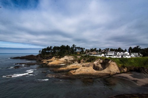 Coastal view featuring rocky cliffs, a sandy beach, and houses nestled among trees under a cloudy sky.