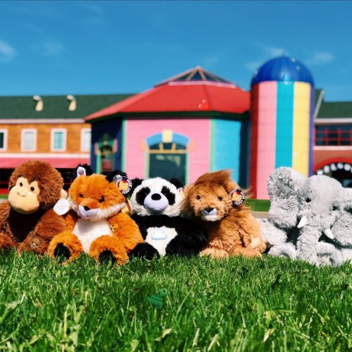 A row of colorful stuffed animals on green grass, with a vibrant building in the background under a clear blue sky.