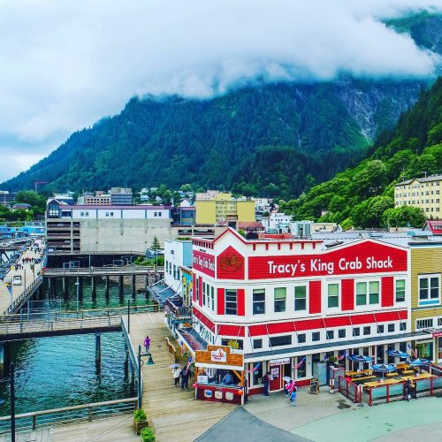 A colorful seafood restaurant by the water, surrounded by mountains and clouds, with people walking nearby.