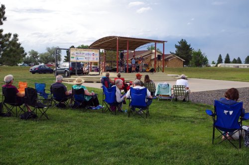 A crowd sits in folding chairs, watching a live band perform on an outdoor stage in a grassy area.