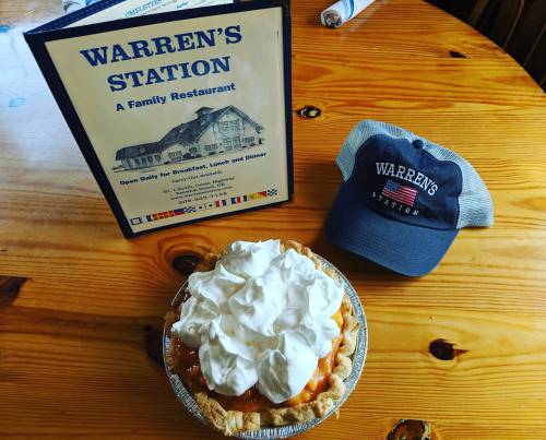 A pie topped with whipped cream sits next to a menu and a cap from Warren's Station restaurant on a wooden table.