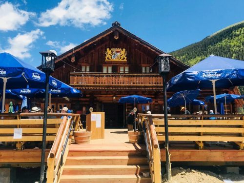 A rustic wooden lodge with blue umbrellas on a sunny day, surrounded by green mountains and people enjoying the outdoors.