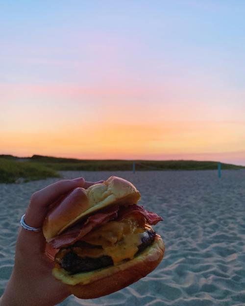 A hand holding a cheeseburger with bacon against a colorful sunset over a sandy beach.