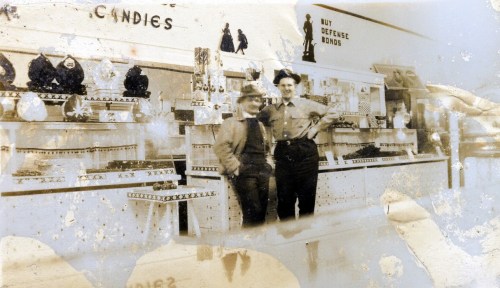 Two men stand in a vintage candy store, surrounded by displays of sweets and war bond advertisements.