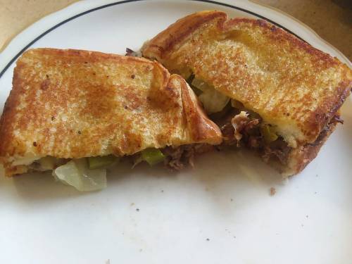 A toasted sandwich cut in half, filled with meat and pickles, served on a white plate with a black rim.
