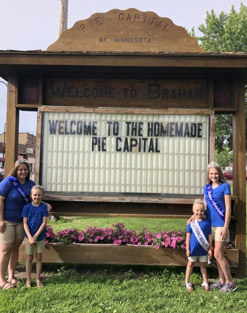 Four people stand in front of a sign that reads "Welcome to the Homemade Pie Capital of Minnesota" in Braham.