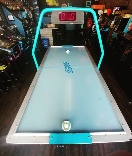 Aerial view of an air hockey table with a digital score display, set in a vibrant arcade environment.