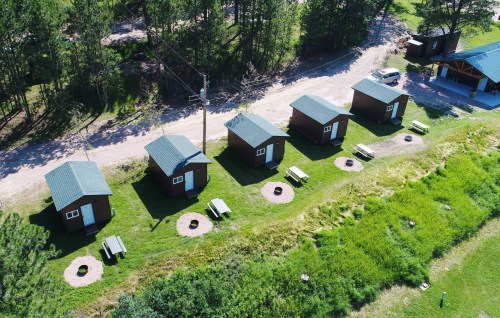Aerial view of five small cabins with picnic tables and fire pits, surrounded by green grass and trees.
