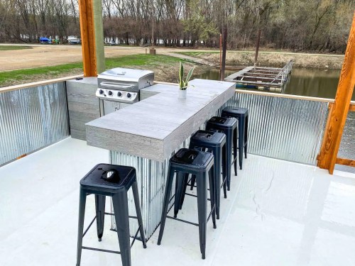 Outdoor bar area featuring a grill, a long table, and black stools, overlooking a serene water view.