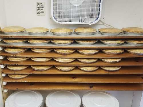 A shelf stacked with multiple pies in metal pans, with a fan mounted above and containers below.