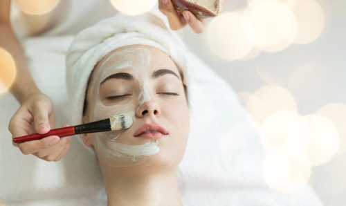 A woman receiving a facial treatment, with a hand applying a mask using a brush, in a serene spa setting.