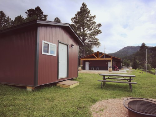A small brown cabin and a larger building with a porch, surrounded by green grass and trees in a scenic outdoor setting.