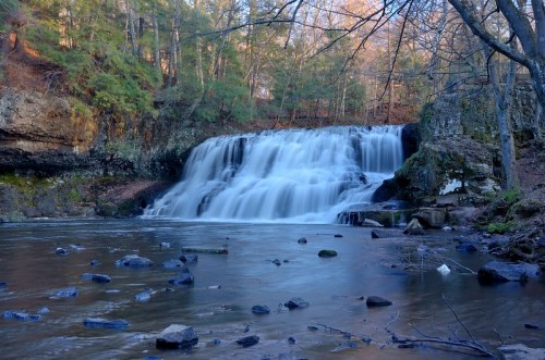 A serene waterfall cascades over rocks, surrounded by trees and a calm river in a tranquil forest setting.