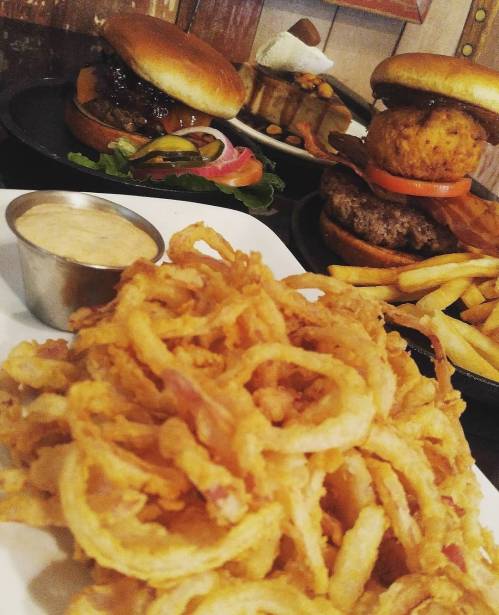 A plate of crispy onion rings in the foreground, with burgers and fries in the background.