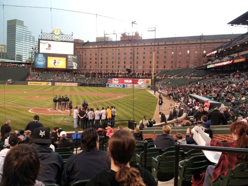 A baseball stadium with fans in the stands, players on the field, and a city skyline in the background.