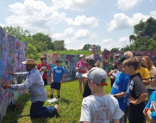 A group of children watches an artist painting a colorful mural on a wall under a sunny sky.