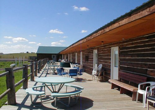A wooden deck with tables and chairs, overlooking a grassy field under a blue sky with scattered clouds.
