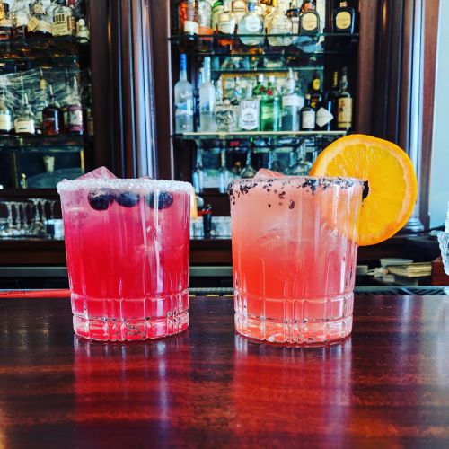 Two colorful cocktails on a wooden bar, one garnished with an orange slice and the other with blueberries and a sugar rim.