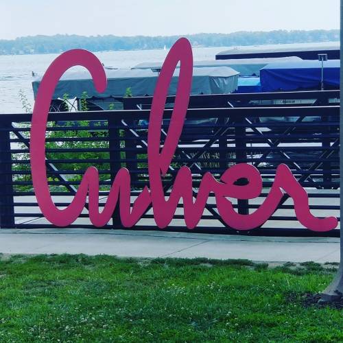 A large, pink sign reading "Culver" stands near a lake with boats in the background and greenery around.