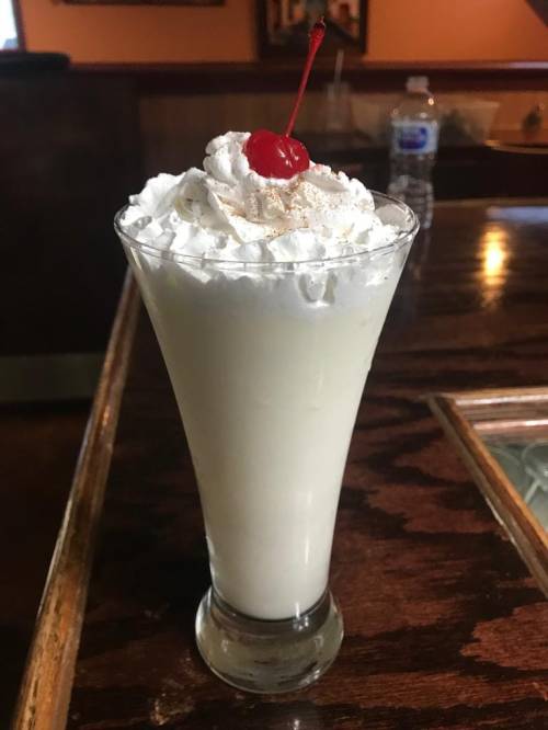 A tall glass of creamy dessert topped with whipped cream and a cherry, sitting on a wooden bar counter.