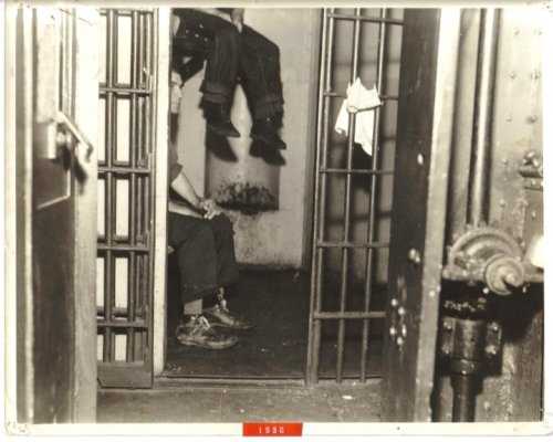 A historic black-and-white photo of a prison cell, showing a seated person and another figure's legs hanging above.