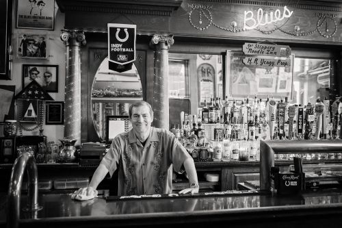 A bartender stands at a vintage bar, surrounded by bottles and memorabilia, in a cozy, nostalgic setting.
