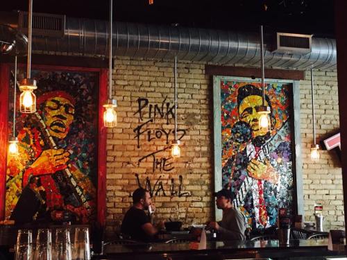 Two patrons sit at a bar with colorful mosaic art of musicians on the wall, illuminated by hanging light fixtures.