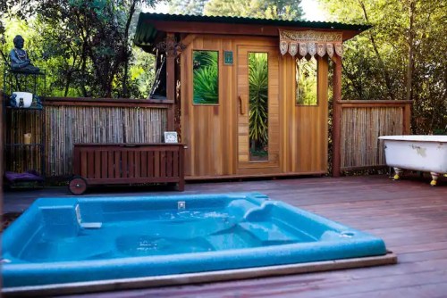 A wooden sauna beside a hot tub on a deck, surrounded by greenery and decorative elements.