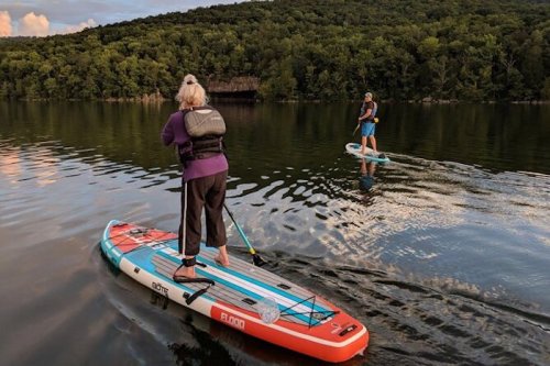 Two people paddleboarding on a calm lake surrounded by lush greenery, with reflections on the water's surface.