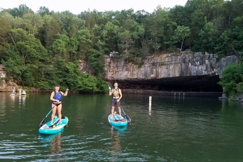 Two people paddleboarding on a calm lake surrounded by lush greenery and a rocky cliff in the background.