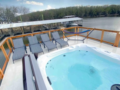 A hot tub on a deck overlooking a river, with several lounge chairs and a hammock nearby.