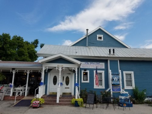 A blue restaurant with a white porch and sign, surrounded by greenery under a partly cloudy sky.