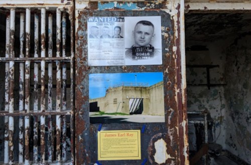 A weathered wall displays FBI wanted posters and information about James Earl Ray, near prison bars.