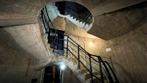 A spiral staircase in a concrete structure, illuminated by a single light, with a dark, industrial atmosphere.