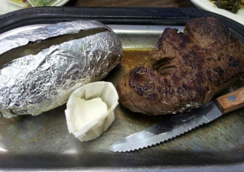 A grilled steak with a baked potato wrapped in foil and a dollop of butter on a metal plate.