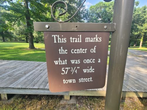 Sign indicating the center of a former 57¾" wide town street, set in a park with trees and a wooden walkway.