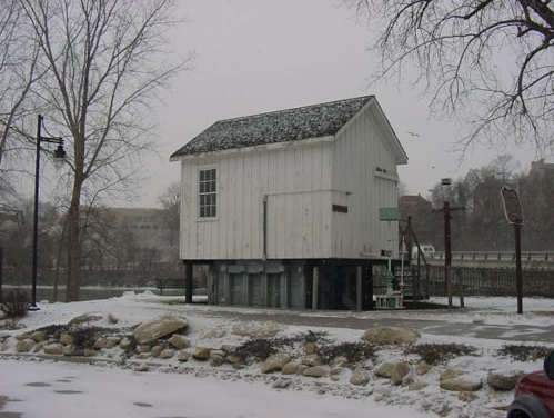 A small white building on stilts in a snowy landscape, surrounded by rocks and bare trees.