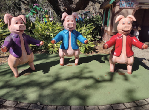 Three cartoonish pig statues in colorful jackets stand on a grassy area, surrounded by greenery.
