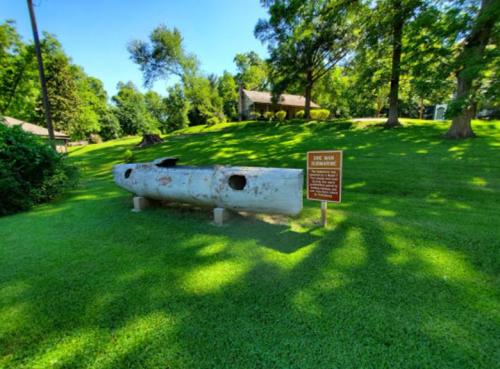 A weathered, old canoe on a grassy area, next to a sign, surrounded by trees and a house in the background.