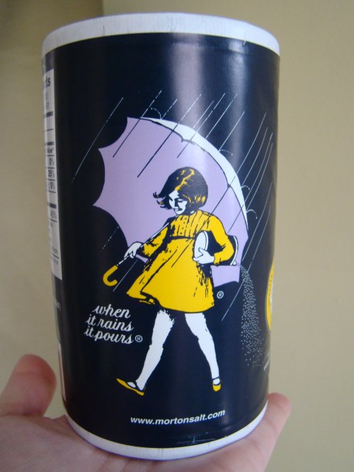 A hand holding a cylindrical container of Morton Salt featuring a girl with an umbrella and the phrase "when it rains it pours."