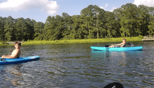 Two young men kayaking on a calm lake surrounded by lush green trees under a blue sky.