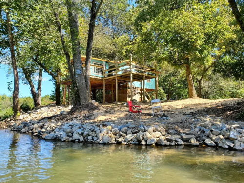 A lakeside view of a wooden house surrounded by trees, with a red chair and a white chair on the shore.